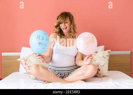 A smiling and happy pregnant caucasian woman sitting in bed holding two balloons. She is teasing with balloons with the inscription 'its a boy' and 'i Stock Photo