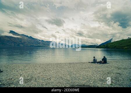 Queenstown New Zealand - February 28 2015; Three young adult travellers relaxing in early morning light on shore of Lake Wakatipu in low light split t Stock Photo