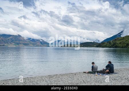 Queenstown New Zealand - February 28 2015; Three young adult travellers relaxing in early morning light on shore of Lake Wakatipu in low light split t Stock Photo