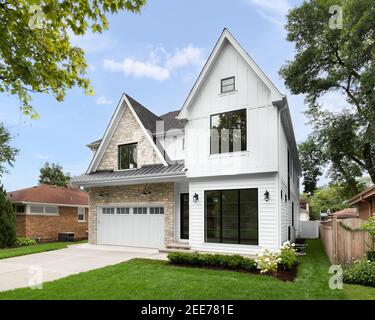 A new, white modern farmhouse with a dark shingled roof and black windows. The left side of the house is covered in a rock siding. Stock Photo