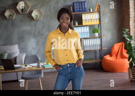 Photo of confident young afro american business woman hold hands pockets wear yellow shirt jeans stand in office indoors Stock Photo