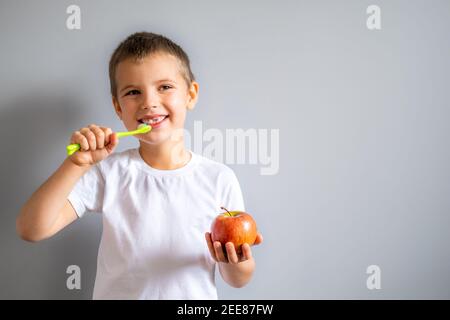 Boy without milk upper tooth in white t-shirt smiling and holding an apple and toothbrush in hands on the gray background. Stock Photo