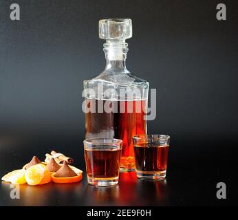 A glass bottle and two glasses of cognac stand on a black background next to sweets, slices and tangerine zest. Close-up. Stock Photo