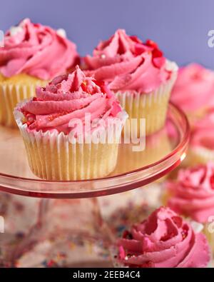 A display of bright pint frosted cupcakes sit on a clear pink cake stand Stock Photo