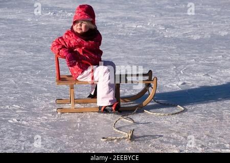 Little girl, wrapped in a red winter coat, hat, gloves and white pants and with skates underneath sits on a sled on the ice Stock Photo