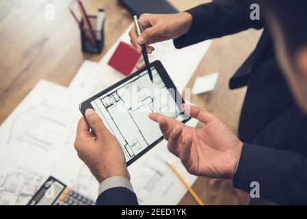 Real estate agent with client or architect team discussing a housing model and its blueprints digitally using a tablet computer Stock Photo