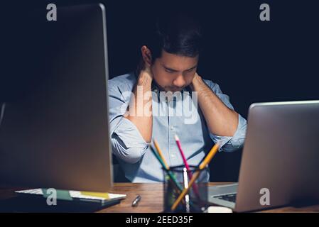 Stressed tired young man having office syndrome , neck pain, while working overtime on his laptop computer at night finishing his project Stock Photo