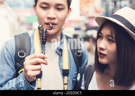 Asian couple tourist backpackers traveling on holidays, holding and looking at fried scorpion one of street food at Khao San road in Bangkok, Thailand Stock Photo