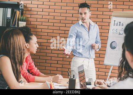 Young handsome hispanic businessman leader making a presentation, sharing ideas and projections with his colleagues in an office team meeting Stock Photo