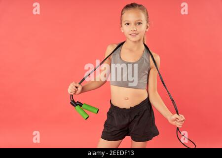 Adorable female child in sportswear looking at camera and smiling while holding jump rope Stock Photo