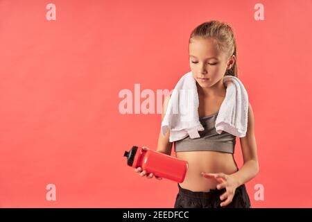 Adorable female child with towel on her shoulders looking at sports drink with serious expression while standing against red background Stock Photo