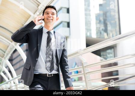Enthusiastic Thai-Asian Business man walking while talking on mobile phone in the city on the way to work at the office Stock Photo