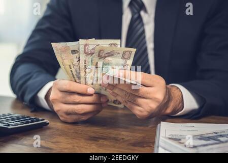 A man counting money, UAE Dirham banknotes, and calculating the total value with a calculator before signing an agreement. Stock Photo