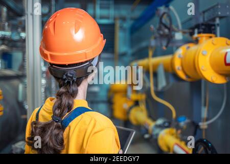 A female engineer in uniform and a protective helmet, holding a tablet and conducting an inspection of the equipment. The view from the back. The conc Stock Photo