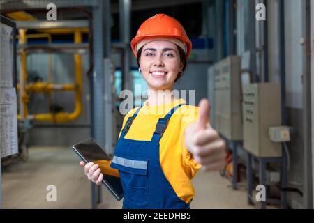 Industrial production. A female engineer in uniform and a protective helmet, holding a tablet in her hands, gives a thumbs up. The concept of equality Stock Photo