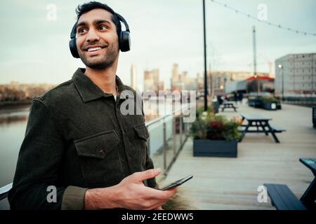 Smiling mixed race male young adult leaning against railing listening to music on smartphone wearing headphones Stock Photo