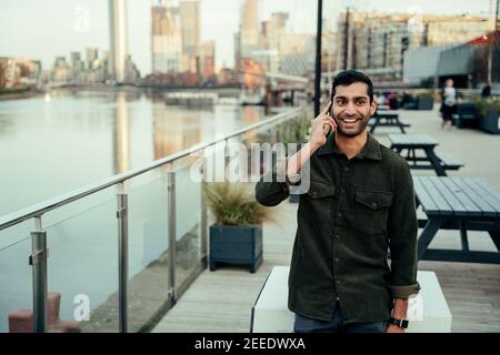 Mixed race male chatting on smartphone walking through harbour smiling Stock Photo