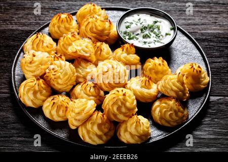 Duchess potatoes, mashed potatoes piped in decorative swirls, browned in an oven served with white sauce on a black plate on a wooden table Stock Photo