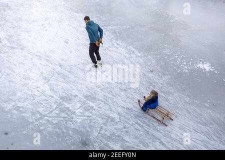 ZUTPHEN, NETHERLANDS - Feb 14, 2021: Father ice skating holding a rope dragging a child on a sled on frozen river seen from above with marks in the ic Stock Photo