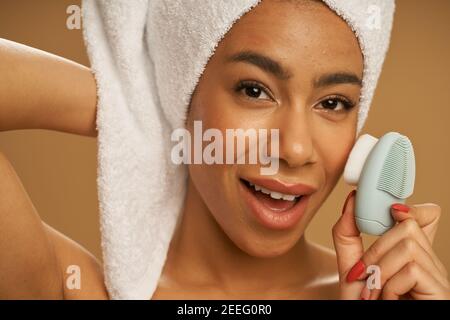 Face closeup of playful young woman looking at camera while using silicone facial cleansing brush, posing isolated over beige background. Skincare concept Stock Photo