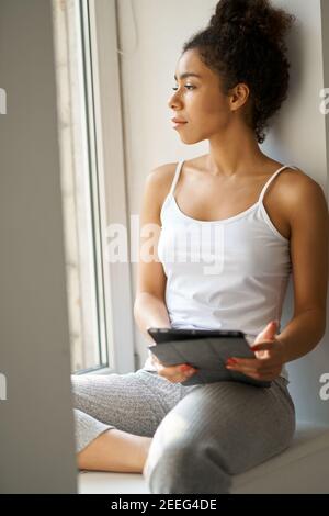 Thoughtful young woman with tablet pc looking out the window, feeling cozy while sitting alone by the window at home. Lifestyle, weekend concept Stock Photo