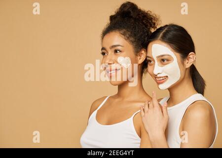 Portrait of two young women, female friends smiling aside while posing together with facial masks on isolated over beige background. Skincare, beauty concept Stock Photo