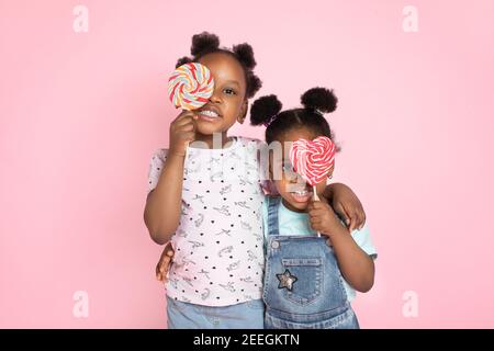 Close up horizontal shot of two cute little African girls covering her eyes with sweet colorful lollipops, smiling to camera on pink color background Stock Photo
