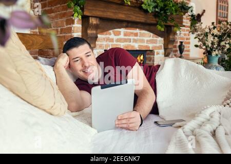 Happy man chatting on digital tablet app while relaxing in cozy bed at home Stock Photo