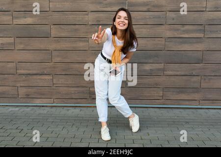 Full length body size photo of pretty brunet young happy woman wearing casual clothes and listening to music via wireless earphones standing in the Stock Photo