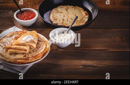 Traditional Russian Crepes Blini stacked in plate and cast-iron frying pan with red caviar, fresh sour cream on dark wooden table. Russian festival me Stock Photo