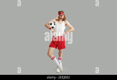 Happy nerdy man in glasses, tank top and shorts standing akimbo, holding soccer ball and smiling Stock Photo
