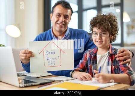Happy latin boy sitting at the desk together with his father, using laptop during remote learning at home. Dad holding Stay home drawing. Online education, homeschooling concept Stock Photo