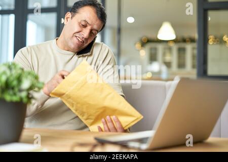 Portrait of hispanic middle aged man talking on the phone while working from home, sitting at the desk with his laptop. Focus on the man. Telework, business concept Stock Photo