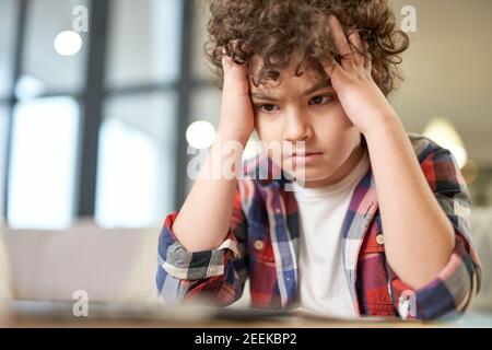 Portrait of tired latin boy holding his head, looking sad while sitting at the desk and studying at home. Online education, homeschooling concept Stock Photo