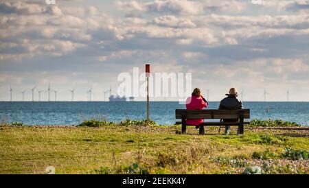 With the Rampion offshore wind farm in the distance a couple sit on a bench looking at the view. Shoreham, West Sussex, UK. Stock Photo