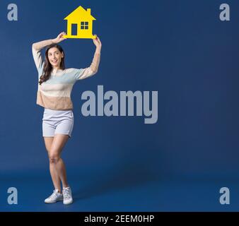 Happy young smiling Asian woman holding and showing yellow color house mockup paper on blue color background, thinking for investment, sales and insur Stock Photo