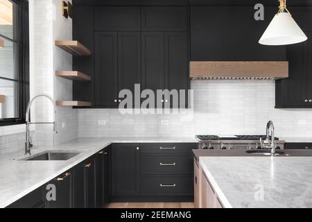 A luxury kitchen with black cabinets, granite counter top, tiled backsplash, stainless steel appliances, and chrome faucets. Stock Photo