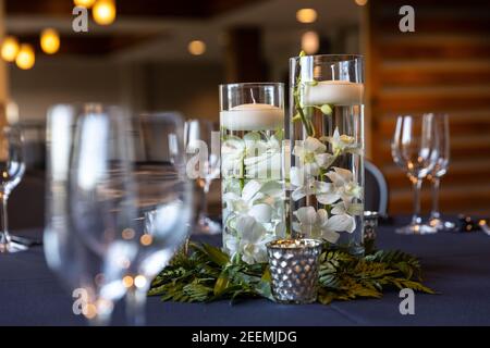 A centerpiece at a wedding with wine glasses on the table, flowers and floating candles in pillar vases, and blurred out lights in the distance. Stock Photo
