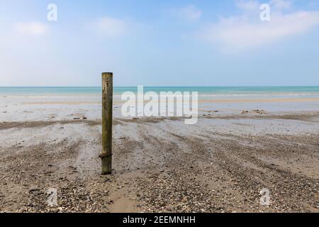 A solitary wooden post on a softly lit pebble strewn beach backed by a calm blue sea and blue sky with soft clouds and copy space Stock Photo