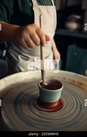 Child makes earthenware cup working on pottery wheel at workshop and paints it using paintbrush. Stock Photo