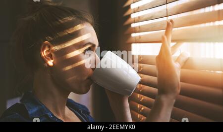 woman looking through window blinds into the sunlight and drinking coffee Stock Photo