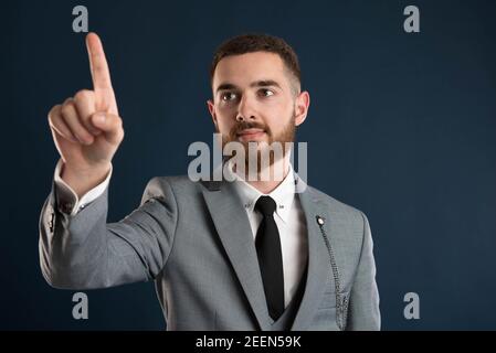 Smiling young businessman pointing at a virtual screen wearing a grey suit and black tie Stock Photo