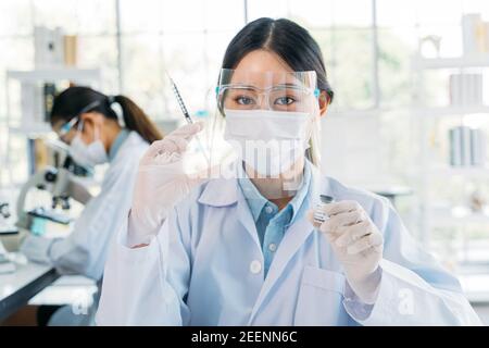 Successful young female medical researcher and scientist wearing labcoat standing in laboratory while working on developing covid-19 vaccine holding injection and bottle Stock Photo