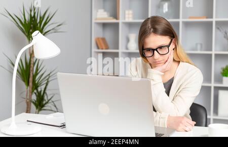 Girl student thoughtfully listens to a lesson online using a laptop. A young woman is looking at the computer screen with concentration. Stock Photo