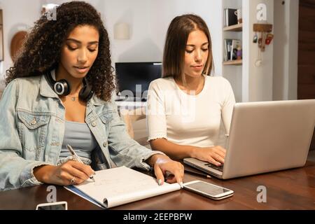 Two young millennials women self entrepreneur working from home with laptop and smartphone in the living room writing notes. New Normal internet jobs Stock Photo