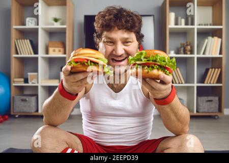 Man sportsman in sportswear sitting and holding two fresh unhealthy burgers in hands Stock Photo