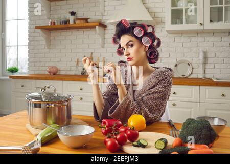 Angry young woman in hair curlers sitting at kitchen table, filing nails and thinking