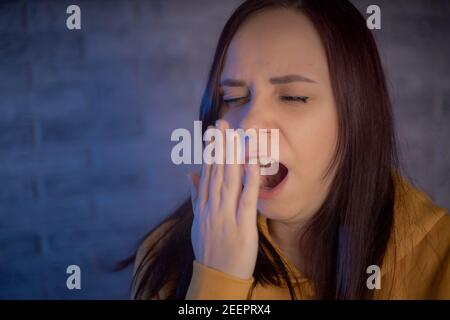 A yawning woman against background of gray brick wall. Young brunette covers her mouth and yawns in the morning Stock Photo