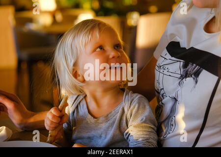 Young blond mother relaxing in sunny indoor cafe text messaging with her phone with her little cute child eating pizza. Stock Photo
