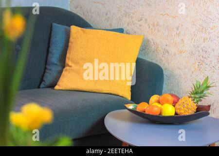 Cozy living room interior, gray sofa and yellow pillows. Plate or basket with different tropical fruits - pineapple, apples, citruses on the little gr Stock Photo